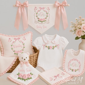 May include: A collection of baby items including a wall banner, a ruffled pillow, a short-sleeve onesie, a bib, a security blanket with a teddy bear head, and a cloth, all featuring pink rose embroidery, bows, and the name Hailey Rose.