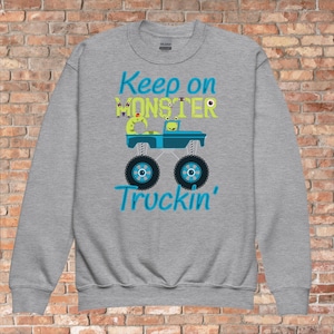 Op de afbeelding: Grijs sweatshirt met een blauwe monstertruck afbeelding en de tekst "Keep on Monster Truckin'" in blauwe en groene letters.