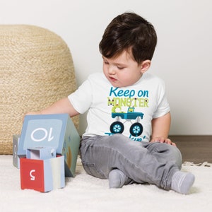 K&ouml;nnte beinhalten: Ein Kleinkind tr&auml;gt ein wei&szlig;es T-Shirt mit einem gr&uuml;nen Monstertruck-Motiv und dem Text "Keep on Monster Truckin'" in Blau und Gr&uuml;n. Das Kleinkind sitzt auf einer wei&szlig;en Oberfl&auml;che und spielt mit Bl&ouml;cken.