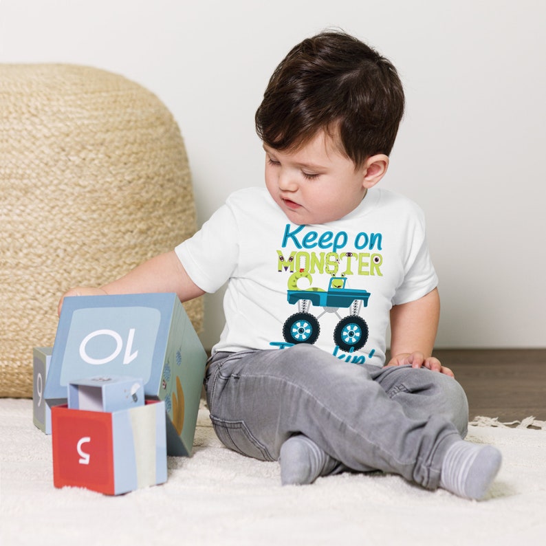 K&ouml;nnte beinhalten: Ein Kleinkind tr&auml;gt ein wei&szlig;es T-Shirt mit einem gr&uuml;nen Monstertruck-Motiv und dem Text "Keep on Monster Truckin'" in Blau und Gr&uuml;n. Das Kleinkind sitzt auf einer wei&szlig;en Oberfl&auml;che und spielt mit Bl&ouml;cken.