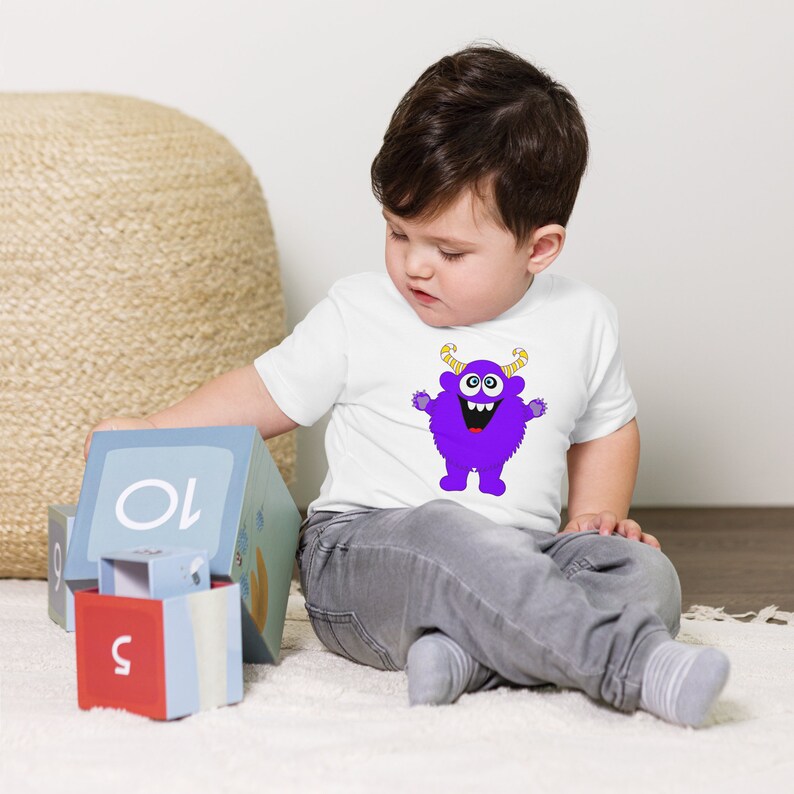 以下が含まれることがあります： 白いTシャツを着た幼児が、紫色のモンスターのグラフィックを着ています。モンスターは黄色い角、大きな目、そして大きな笑顔をしています。幼児は白い敷物に座って、カラフルなブロックで遊んでいます。