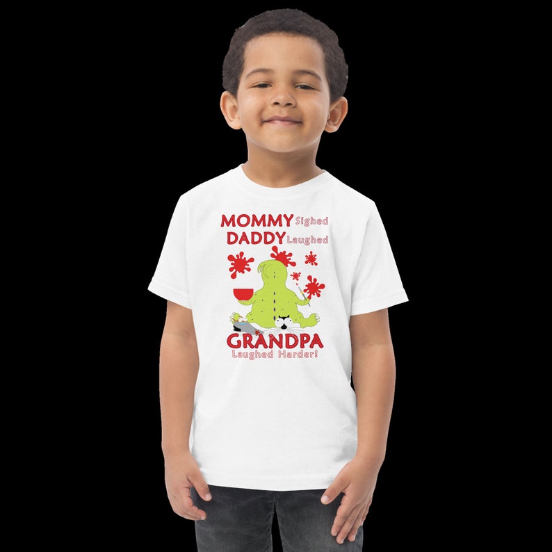 Puede incluir: Camiseta blanca con un personaje de dibujos animados verde pintando con salpicaduras de pintura roja. El texto en la camiseta dice "Mam&aacute; suspir&oacute;, pap&aacute; se ri&oacute;, &iexcl;abuelo se ri&oacute; m&aacute;s fuerte!"