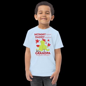 Puede incluir: Una camiseta azul claro con un personaje de dibujos animados verde que sostiene un pincel y un taz&oacute;n rojo. El texto en la camiseta dice "Mommy sighed, Daddy laughed, Grandpa laughed harder!"