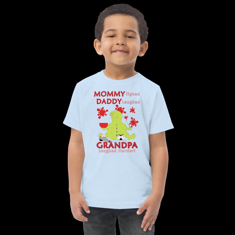 Puede incluir: Una camiseta azul claro con un personaje de dibujos animados verde que sostiene un pincel y un taz&oacute;n rojo. El texto en la camiseta dice "Mommy sighed, Daddy laughed, Grandpa laughed harder!"