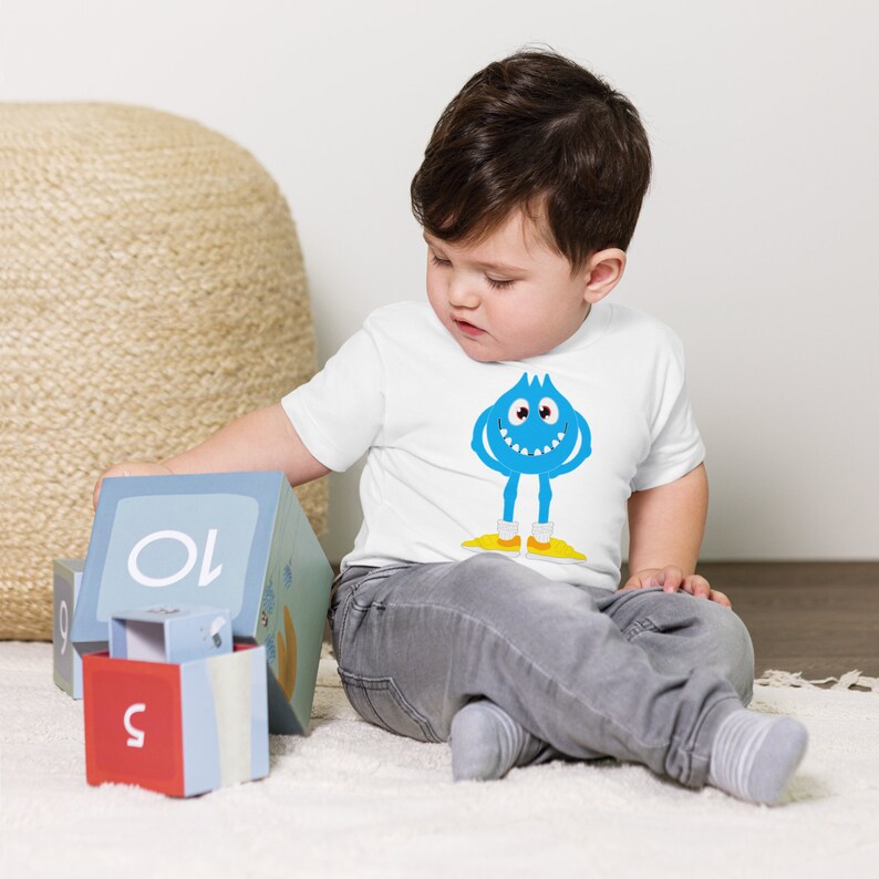 K&ouml;nnte beinhalten: Ein Kleinkind tr&auml;gt ein wei&szlig;es T-Shirt mit einem blauen Monster-Motiv. Das Monster hat ein zahniges Grinsen und gelbe Schuhe. Das Kleinkind sitzt auf einem wei&szlig;en Teppich und spielt mit bunten Bl&ouml;cken.