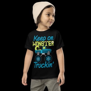 K&ouml;nnte beinhalten: Ein schwarzes T-Shirt mit einem blauen und gr&uuml;nen Monstertruck-Motiv und dem Text "Keep on Monster Truckin'" darauf. Ein kleines Kind tr&auml;gt das Shirt.