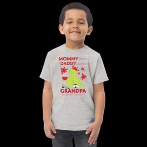 Puede incluir: Una camiseta gris claro con un monstruo verde de dibujos animados que lleva un sombrero rojo y sostiene una taza roja. El texto de la camiseta dice "Mommy Sighed, Daddy Laughed, Grandpa Laughed Harder!"