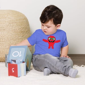May include: A young child wearing a blue t-shirt with a red monster graphic. The monster is wearing glasses and has a big smile. The child is playing with blocks that have numbers on them.