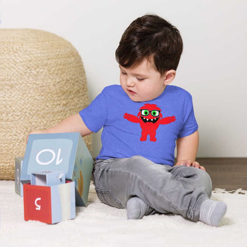 May include: A young child wearing a blue t-shirt with a red monster graphic. The monster is wearing glasses and has a big smile. The child is playing with blocks that have numbers on them.