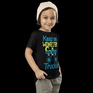 K&ouml;nnte beinhalten: Ein schwarzes T-Shirt mit einem blauen und gr&uuml;nen Monstertruck-Motiv und dem Text "Keep on Monster Truckin'" in blauen und gelben Buchstaben.