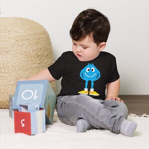K&ouml;nnte beinhalten: Ein Kleinkind tr&auml;gt ein schwarzes T-Shirt mit einem blauen Monster-Motiv. Das Monster hat ein breites L&auml;cheln und gelbe Schuhe. Das Kleinkind spielt mit bunten Bl&ouml;cken.