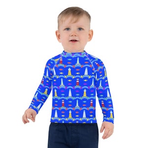 May include: A blue long-sleeved rash guard for kids with a pattern of lighthouses, waves, and fish. The rash guard has a mock neck and long sleeves.