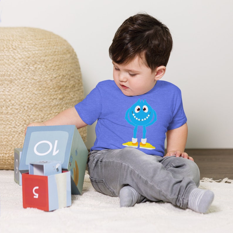 K&ouml;nnte beinhalten: Ein kleines Kind tr&auml;gt ein blaues T-Shirt mit einem blauen Monster-Motiv. Das Monster hat ein breites L&auml;cheln und gelbe Schuhe. Das Kind spielt mit bunten Bl&ouml;cken, auf denen Zahlen stehen.