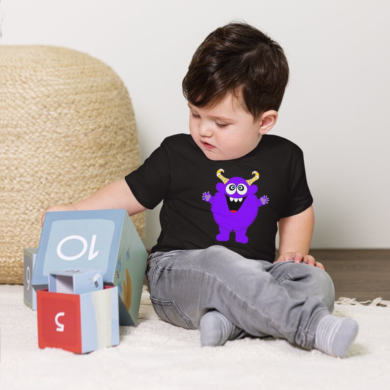 以下が含まれることがあります： 黒いTシャツを着た幼児が、紫色のモンスターのグラフィックを着ています。モンスターは黄色い角、大きな目、そして大きな笑顔をしています。幼児は白い敷物に座って、カラフルなブロックで遊んでいます。
