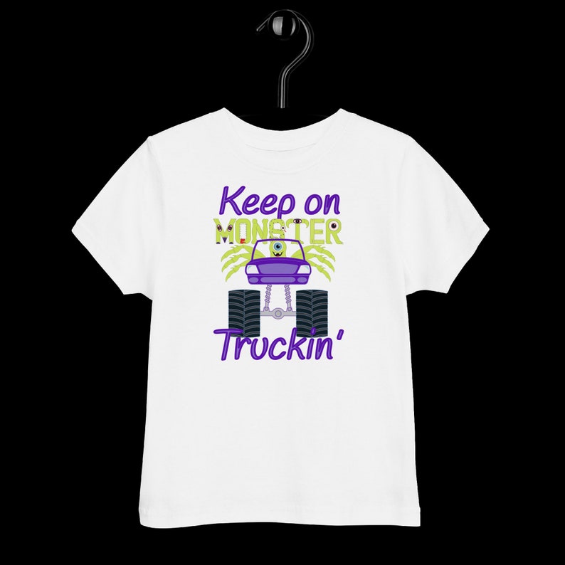 May include: White toddler t-shirt with a green monster truck graphic. The truck has purple wheels and a purple monster face with one eye. The text "Keep on Monster Truckin'" is printed in purple on the shirt.