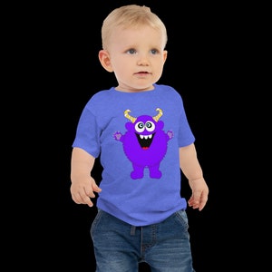 以下が含まれることがあります： 青いTシャツを着た幼児。Tシャツには紫色のモンスターのプリントがされています。モンスターは黄色い角と大きな目、そして大きく笑っています。