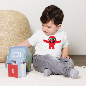 May include: A young child wearing a white t-shirt with a red monster graphic wearing glasses. The monster has green eyes and a toothy grin. The child is playing with blocks that have numbers on them.