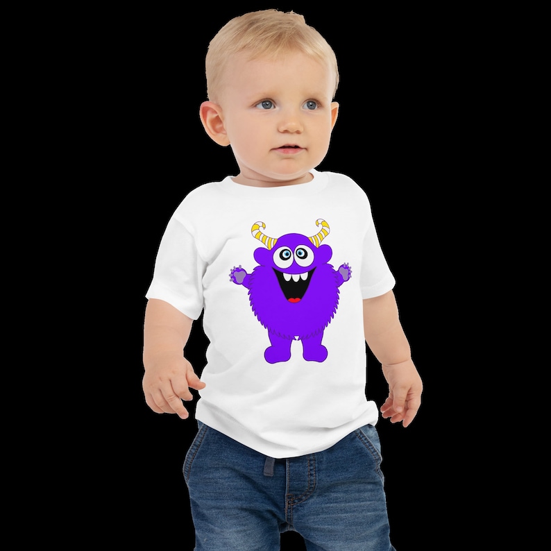 以下が含まれることがあります： 紫色のモンスターのグラフィックがプリントされた白いベビーTシャツ。モンスターは黄色い角、大きな目、そして大きく笑っています。