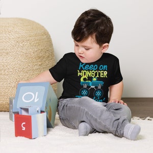 K&ouml;nnte beinhalten: Ein kleines Kind tr&auml;gt ein schwarzes T-Shirt mit dem Text "Keep on Monster Truckin'" in gelben und blauen Buchstaben. Das Shirt zeigt einen Monstertruck aus der Zeichentrickserie mit einem gr&uuml;nen Monsterfahrer. Das Kind sitzt auf einem wei&szlig;en Teppich und spielt mit Bl&ouml;cken.