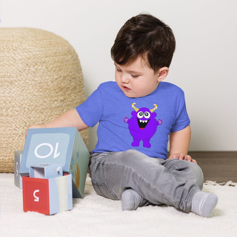 以下が含まれることがあります： 青いTシャツを着た幼児が、紫色のモンスターのグラフィックを描いています。モンスターは黄色い角、大きな目、そして大きな笑顔をしています。幼児はカラフルなブロックで遊んでいます。