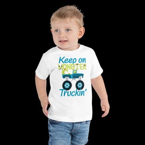 K&ouml;nnte beinhalten: Wei&szlig;es T-Shirt f&uuml;r Kleinkinder mit einem blauen Monstertruck-Motiv und dem Text "Keep on Monster Truckin'" in gr&uuml;ner und blauer Schrift.