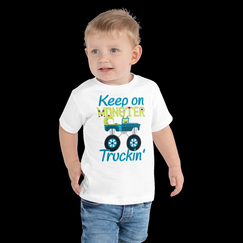 K&ouml;nnte beinhalten: Wei&szlig;es T-Shirt f&uuml;r Kleinkinder mit einem blauen Monstertruck-Motiv und dem Text "Keep on Monster Truckin'" in gr&uuml;ner und blauer Schrift.