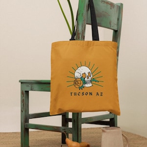 May include: A mustard yellow tote bag with black straps, featuring a white skull with a green rose and the text "TUCSON AZ". The bag is sitting on a green wooden chair with a white flower in the background.