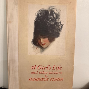 May include: A hardcover book titled A Girl's Life and other pictures by HARRISON FISHER. The cover features a central illustration of a person wearing a large, dark, feathered hat, with red text printed below the artwork on a beige background.