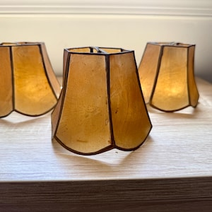 May include: Three amber-colored lamp shades with a translucent, textured appearance. Each shade features a bell-shaped silhouette with a scalloped bottom edge and dark, thin metal framing along the vertical seams and the top and bottom rims.