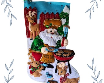Op de afbeelding: Een handgemaakte kerstsok met de kerstman die aan het grillen is. De sok is gemaakt van vilt en versierd met rendieren, een konijn en de tekst "Kiss the Claus". De achtergrond is blauw en wit, met een rode hek en een groene kerstboom.