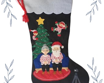 May include: Black Christmas stocking with felt appliqués. Features Santa Claus and a woman wearing matching sweaters, sitting on a blue sofa. A Christmas tree, star, and other festive characters are also included. The top is white with a red hanging loop.