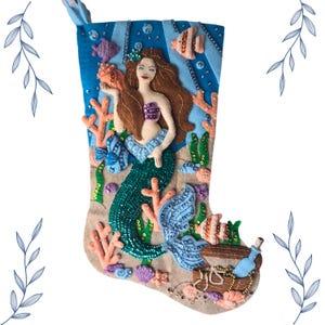 Puede incluir: Un calcetín navideño hecho a mano con un diseño de sirena. El calcetín tiene una cola verde azulado, cabello castaño y una parte superior púrpura. El fondo incluye coral, peces y un cofre del tesoro. El calcetín está decorado con cuentas y lentejuelas.