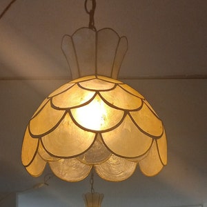 Può includere: Un lampadario con un grande paralume a conchiglia in una calda tonalità gialla. L'apparecchio ha un paralume più piccolo e abbinato sotto. La luce è accesa, illuminando la stanza. L'apparecchio è sospeso al soffitto da una catena.