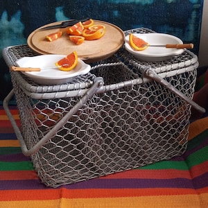 May include: A vintage wire mesh picnic basket with a hinged handle, holding white dishes with orange fruit slices. A wooden cutting board with a knife and more fruit sits atop the basket. The background features a colorful striped textile and a dark blue abstract painting.
