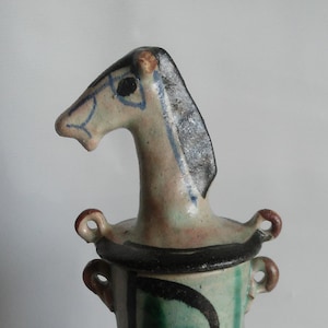 Puede incluir: Un jarrón de cerámica con un diseño de cabeza de caballo. El jarrón es principalmente verde con detalles en negro y blanco. La cabeza del caballo es de color beige claro con detalles azules y negros. El jarrón tiene dos asas pequeñas y un borde negro.