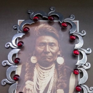 Op de afbeelding: Een set van een zilverkleurige metalen ketting en bijpassende armband met een herhalend krulontwerp. Elk sieraad is versierd met kleine, ronde, dieprode cabochonstenen. De ketting vormt een ovale vorm rondom een vintage portretfoto.