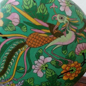 May include: A decorative, oval-shaped cloisonné vase with a vibrant green background. The vase features a detailed design of a bird with pink, yellow, and orange feathers, surrounded by flowers and foliage. The artwork is intricate and colorful.