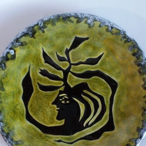 May include: Round ceramic plate with a yellow and green background. A black silhouette of a face and tree-like design is in the center. The rim has a textured, dark gray and white border. Decorative art.