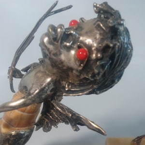 May include: A silver sculpture of a mythical figure with red eyes and a seashell body. The figure is holding a bow and arrow. The sculpture is mounted on a rectangular stone base.