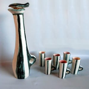 Könnte beinhalten: Ein Keramik-Getränkeset, bestehend aus einer hohen Kanne mit einem fischförmigen Verschluss und sechs passenden Tassen. Die Kanne und die Tassen sind weiß mit vertikalen, teal-farbenen Streifen. Die Tassen haben kleine Henkel und rosa Innenräume.