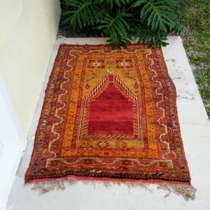 May include: A rectangular, hand-woven rug with a central red field and an arched design. The rug features a border of orange, gold, and brown hues, with intricate patterns. Tassels adorn the bottom edge. The rug is placed on a concrete surface.