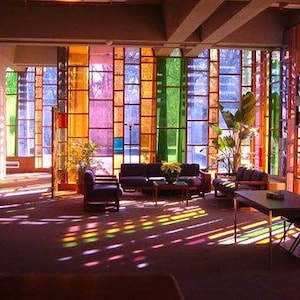 May include: An interior space with large, colorful stained-glass windows. The windows are divided into rectangular sections of orange, yellow, green, and purple. Sunlight streams through, casting colored light on the floor and furniture, including sofas, chairs, and a table.