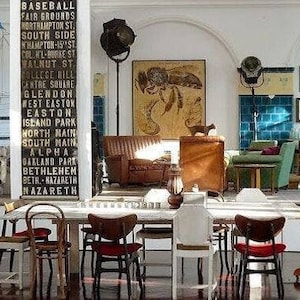 May include: A dining room featuring a long table surrounded by various wooden chairs, a tall pillar with a list of street names, a brown leather sofa, a green armchair, a large bee illustration, and a wall display of multiple mirrors.