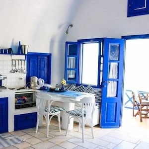 Può includere: Una cucina luminosa con pareti bianche e dettagli blu. Presenta mobili blu, un tavolo bianco con sedie a righe e porte blu aperte. Lo spazio ha uno stile mediterraneo.