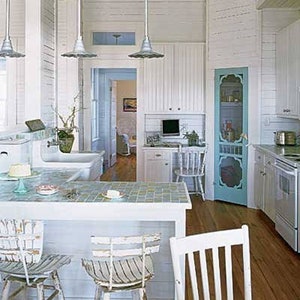 Pode incluir: Uma cozinha clara e arejada com paredes, teto e armários brancos. Possui uma bancada de azulejos, luminárias penduradas e uma porta de tela turquesa. Pisos de madeira e cadeiras brancas completam o design costeiro.