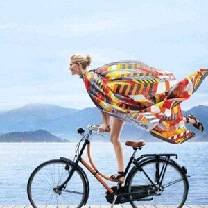 K&ouml;nnte beinhalten: Ein schwarzes und orangefarbenes Fahrrad mit braunem Sattel und Lenker auf einem Holzsteg. Ein gro&szlig;es, wehendes Tuch mit geometrischem Muster in Rot, Gelb, Orange und Schwarz ist dahinter drapiert. Der Text lautet HERMES PARIS LE GOUT DU SPORT.