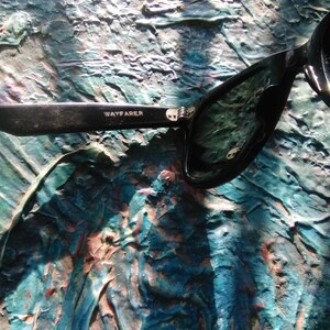 K&ouml;nnte beinhalten: Schwarze Sonnenbrille mit gl&auml;nzendem Finish und dem Wort WAYFARER in wei&szlig;em Aufdruck auf dem inneren B&uuml;gel. Der Rahmen hat eine klassische Form mit einem sichtbaren Metallscharnier am B&uuml;gelansatz und dunkel get&ouml;nten Gl&auml;sern.