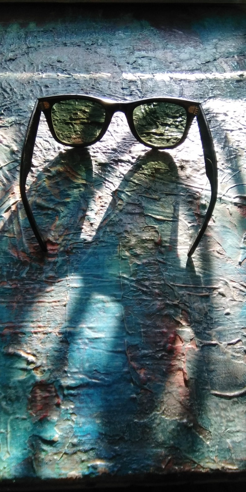 K&ouml;nnte beinhalten: Sonnenbrille mit schwarzem Rahmen und dunkelgr&uuml;n get&ouml;nten Gl&auml;sern. Das Gestell hat eine klassische rechteckige Form mit abgerundeten Kanten und kleinen metallischen Akzenten an den oberen Ecken der Vorderseite.