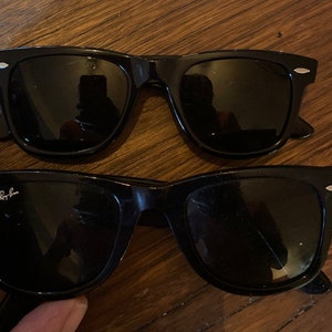 K&ouml;nnte beinhalten: Zwei Paar schwarze Sonnenbrillen mit dunklen Gl&auml;sern und silberfarbenen Akzenten an den Ecken der Rahmen. Ein Paar zeigt das wei&szlig;e Ray-Ban-Logo auf der oberen Ecke des linken Glases. Die Rahmen haben eine klassische rechteckige Form.