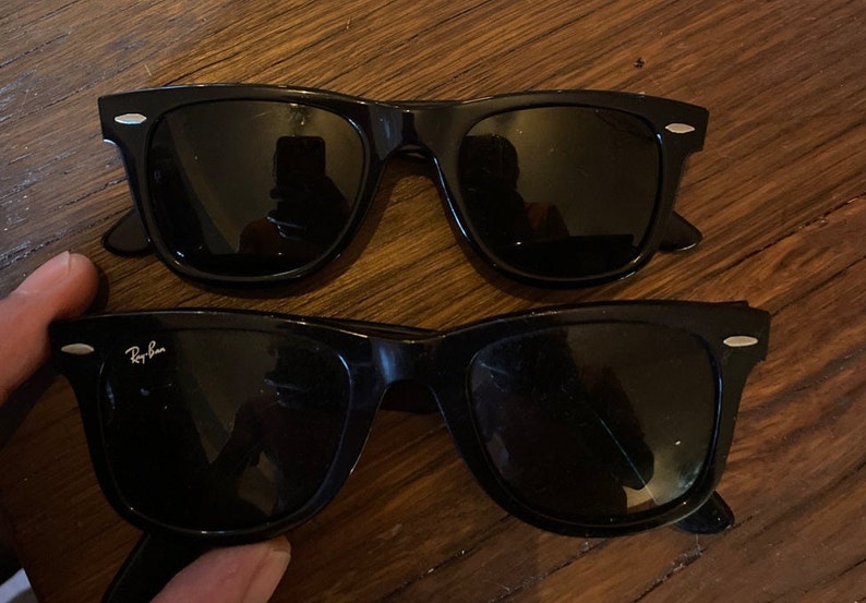 K&ouml;nnte beinhalten: Zwei Paar schwarze Sonnenbrillen mit dunklen Gl&auml;sern und silberfarbenen Akzenten an den Ecken der Rahmen. Ein Paar zeigt das wei&szlig;e Ray-Ban-Logo auf der oberen Ecke des linken Glases. Die Rahmen haben eine klassische rechteckige Form.